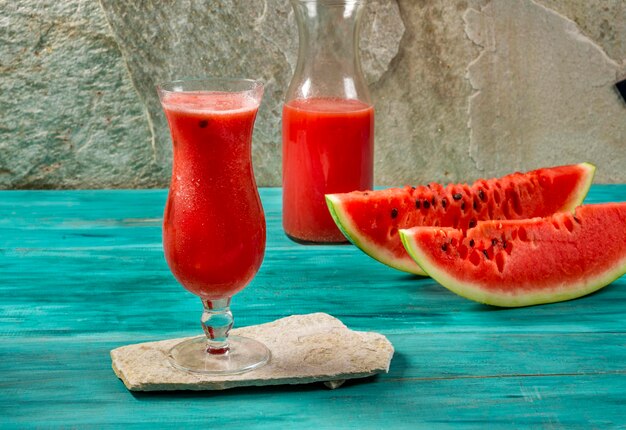 Watermelon juice in cup, watermelon sliced and bottle over blue wood and stone background