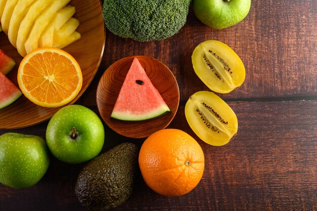 Watermelon orange pineapple kiwi cut into slices with apples and broccoli on a wooden plate and wooden table