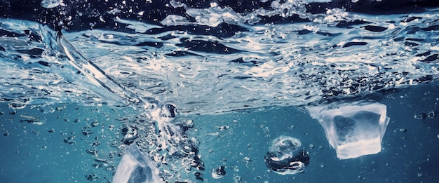 Waterspatten en ijsblokjes Ijs spatten in een glas water Onderwater stromende ijsblokjes die in een heldere waterachtergrond vallen Verfrissende kou drinken Ijs in een glas met blauwe achtergrond