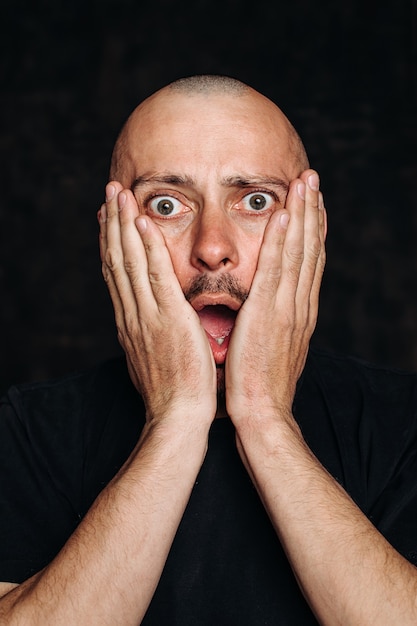 Wauw. Verschrikking op zijn gezicht. Shock, angst. met open mond. Portret van een man in een zwart t-shirt op een zwarte achtergrond, zijn gezicht bedekt met zijn handen. Menselijke emoties, het concept van gezichtsuitdrukking.