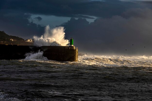 Wave exploding against a lighthouse illuminated by a halo of sunlight about to set Galicia Spain