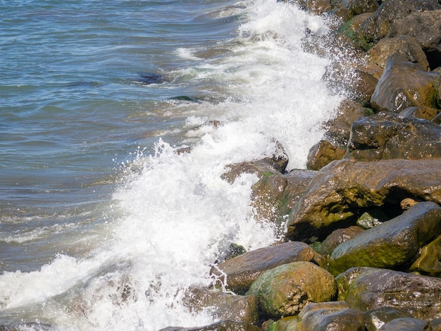 Волны с брызгами падают на большие камни Вид на берег Красивые камни, укрепляющие берег от прибоя Мощная береговая линия Морская пена Волнение моря
