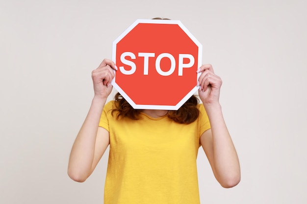 Way prohibited Portrait of anonymous girl in yellow casual Tshirt covering face with stop symbol holding red traffic sign forbidden access Indoor studio shot isolated on gray background