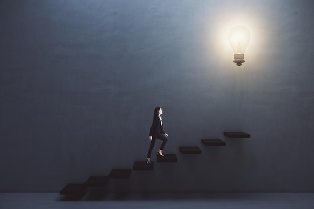 Way to success and creative idea concept with young woman\
climbing the stairs to big illuminated light bulb on dark grey\
wall