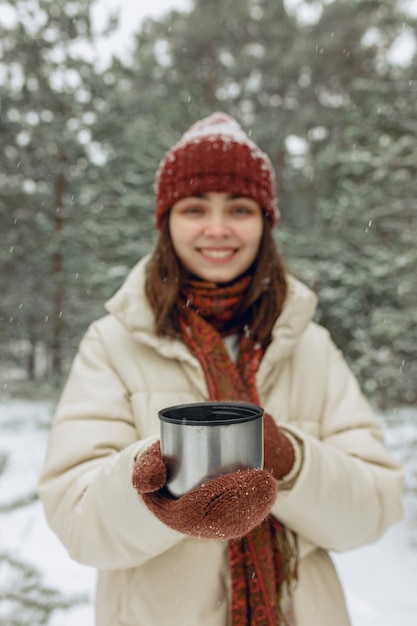 Wazige vrouw met thermoskan kop hete thee die in de winter in het besneeuwde bos staat en handen verwarmt