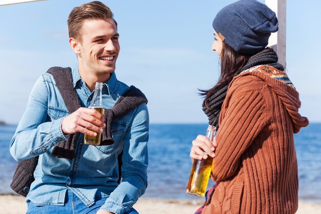 Siamo buoni amici. bel giovane uomo e donna che parlano tra loro e sorridono mentre bevono birra sulla spiaggia