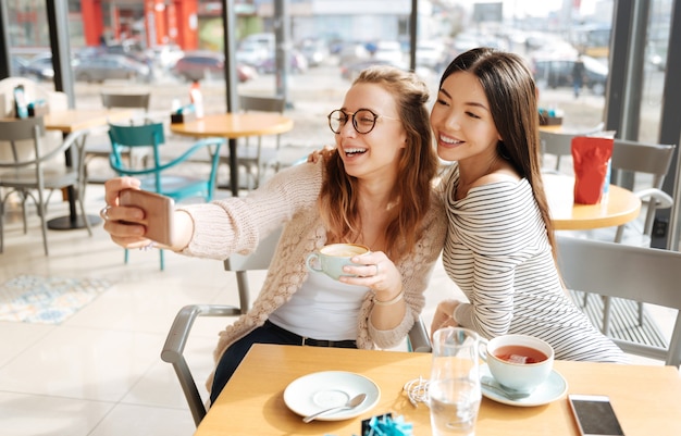 We are happy. Nice portrait of two young girls smiling and taking picture together while sitting at cafe.