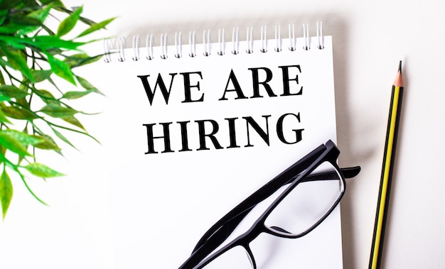 WE ARE HIRING is written in a white notebook next to a pencil, black-framed glasses and a green plant