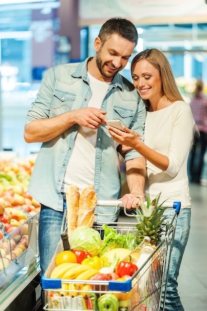 We bought all we need. Happy young couple looking at mobile phone together while standing near shopping cart in food store