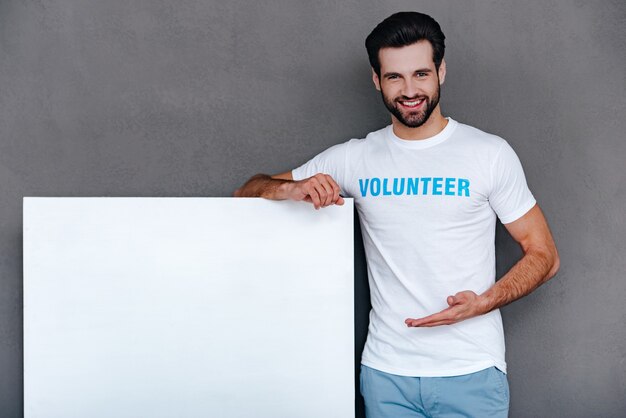 We can help to community! Confident young man in volunteer t-shirt pointing on white boardand looking at camera with smile while standing against grey background