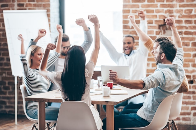 やりました！オフィスのテーブルに座って腕を伸ばして笑顔を保つ6人の陽気な若者のグループ