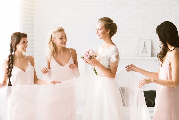 We happy together. Delighted smiling young bridesmaids standing in the white bedroom while helping the bride to get ready and expressing happiness