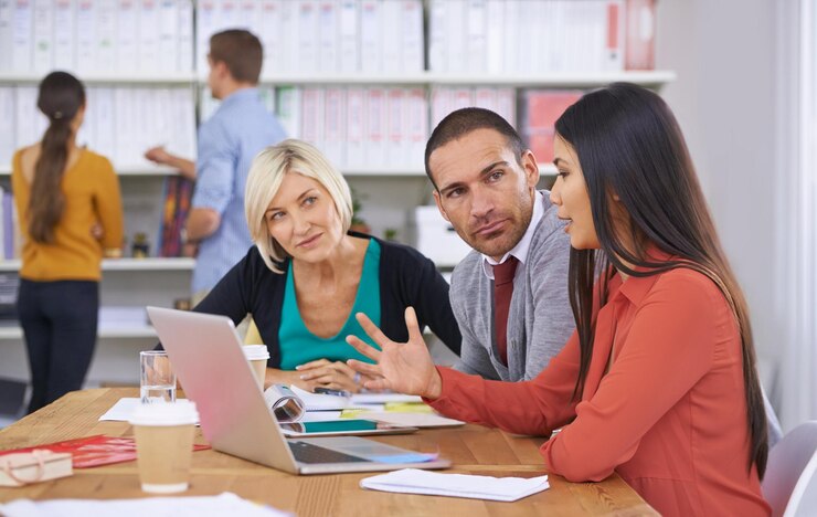 Photo we have to come up with a solution three coworkers brainstorming around a laptop while their colleagues read files in the background