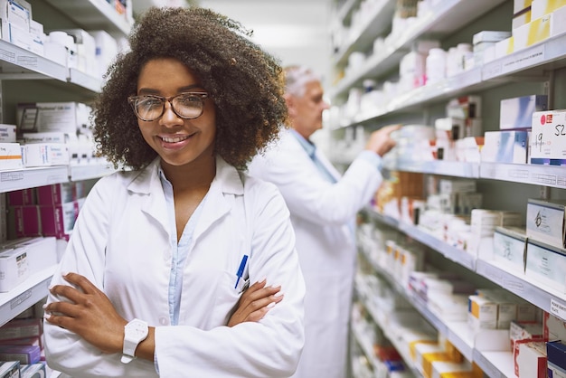 We hebben hier wat je nodig hebt Portret van een vrolijke jonge vrouwelijke apotheker die met gevouwen armen naar de camera in een apotheek kijkt