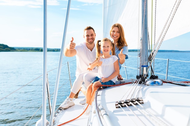 Нам нравится яхтинг. Семья жестикулирует большими пальцами, сидя на палубе парусника, плывущей по морю снаружи, улыбаясь в камеру.