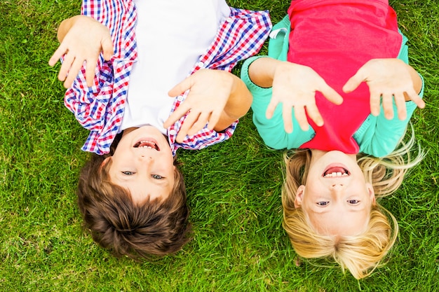 We love summer! Top view of two cute little children raising their hands up and smiling while lying on the green grass together