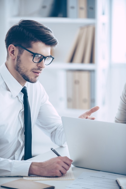 We need to do something with this rates. Close-up part of young handsome man in glasses pointing at laptop while sitting at the office table with his coworker