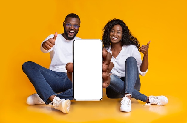 We recommend happy black couple demonstrating smartphone with white blank screen and showing thumb