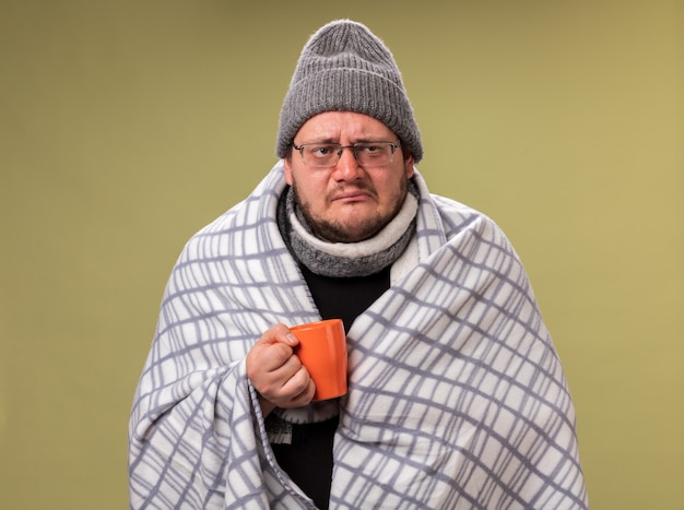 Debole guardando lato maschio malato di mezza età che indossa cappello invernale e sciarpa avvolta in plaid tenendo tazza di tè