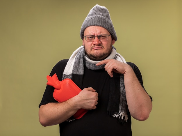 Weak middle-aged ill male wearing winter hat and scarf holding and points at hot water bag isolated on olive green wall