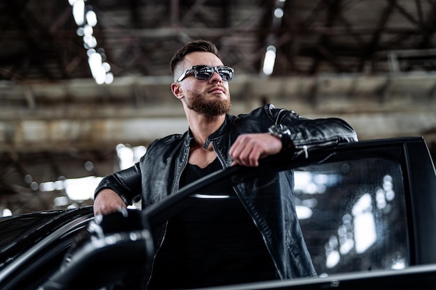 Wealthy life of young man. Cool brutal man near luxury black brend car. Total leather black outfit on man.