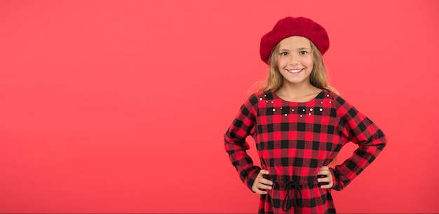 Wear beret like fashion girl. Kid little cute girl with long blonde hair posing in beret hat and checkered dress red background. Beret style inspiration. Fashionable beret accessory for female.