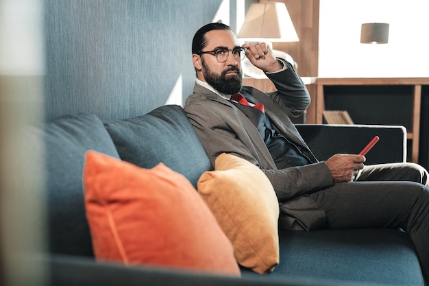 Wear costume. Dark-haired bearded businessman wearing costume sitting on sofa having some rest