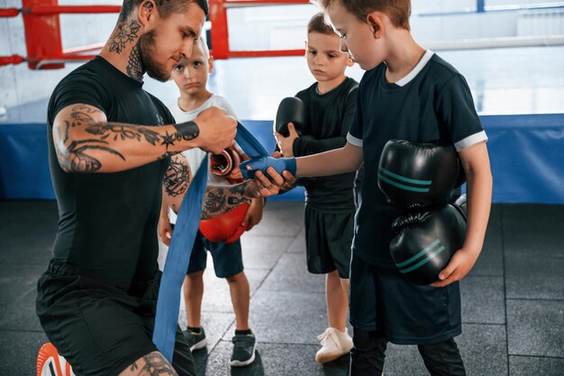 Foto indossare fasce blu per le mani giovane allenatore tatuato che insegna ai bambini le tecniche di boxe