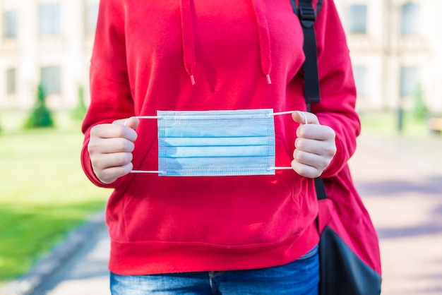 Wearing facial filter blue surgical mask to university concept. Cropped close up photo of young girl in red sweatshirt demonstrating mask