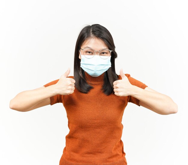 Носить медицинскую маску для предотвращения вируса короны и показывать большие пальцы красивой азиатской женщины, изолированной на белом фоне