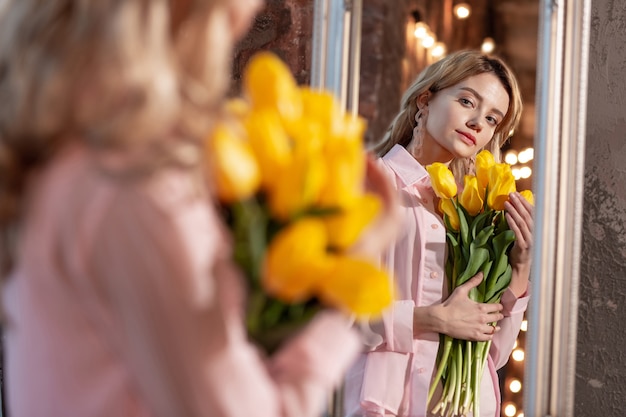 В розовой блузке. Привлекательная женщина в розовой блузке держит желтые цветы и смотрит в зеркало