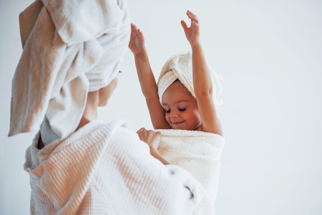 Wearing whiite bathrobes. Young mother with her daugher have beauty day indoors in white room.