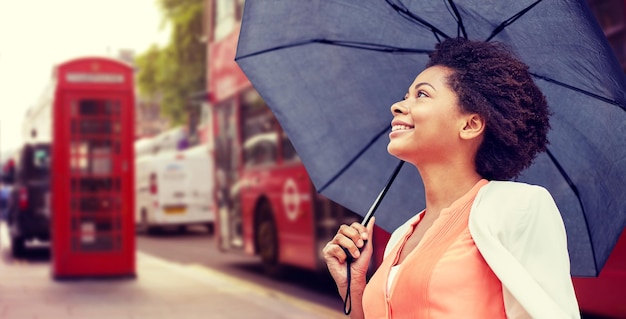 weather, travel, tourism, citylife and people concept - young smiling african american woman with umbrella over london city street background