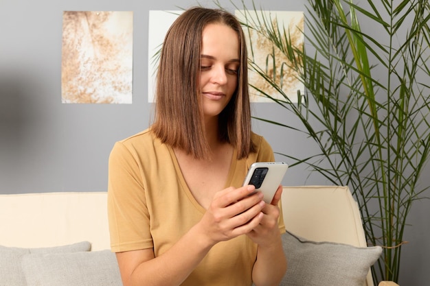 Webbrowsen op smartphone Sociale media-profielen online controleren Freelance werk via smartphone-apps Zelfverzekerde bruinharige vrouw, gekleed in beige T-shirt met telefoon in interieur