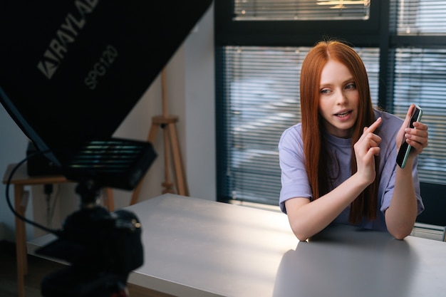 webcam view of smiling young woman blogger greeting followers and talking about phone during video chat on background of window. pov of cheerful redhead female having presentation during sundown.