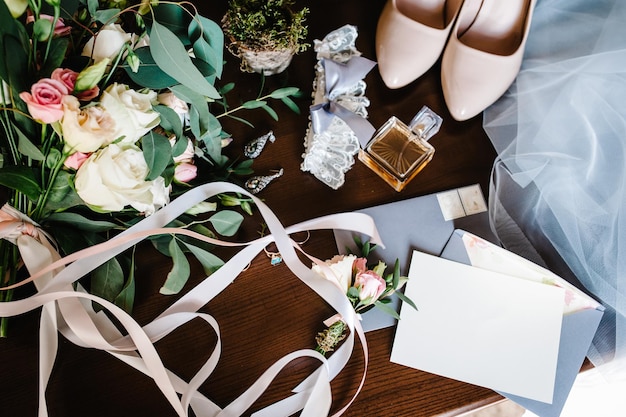 Wedding accessory bride Stylish beige shoes earrings gold rings flowers garter perfumes on table standing on wooden background Letters from the bride and groom flat lay top view