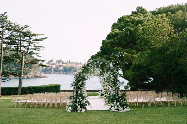 海の近くの緑の芝生の椅子の列に囲まれた結婚式のアーチ