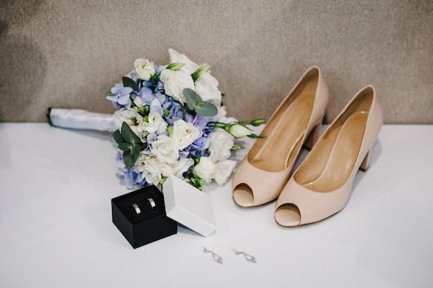 Wedding bouquet of the bride of flowers and greens stylish elegant classic lacquered beige shoes silver earrings and two gold wedding rings lying on pastel background Close up