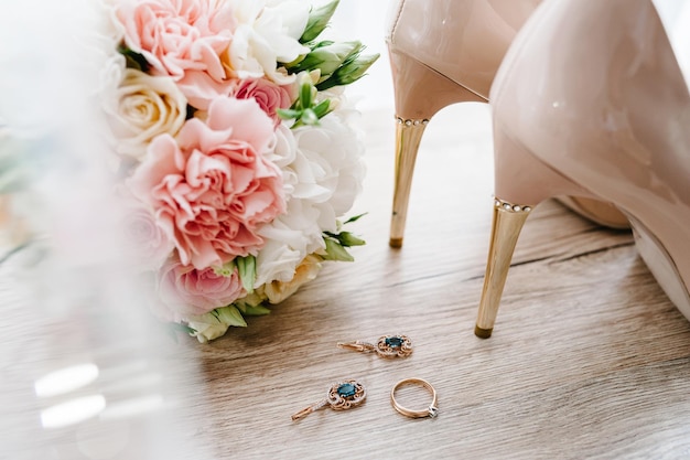 Wedding bouquet of the bride of pink flowers roses and greenery stylish classic lacquered beige shoes earring and ring lying on wooden background Bride accessories Close up Side view