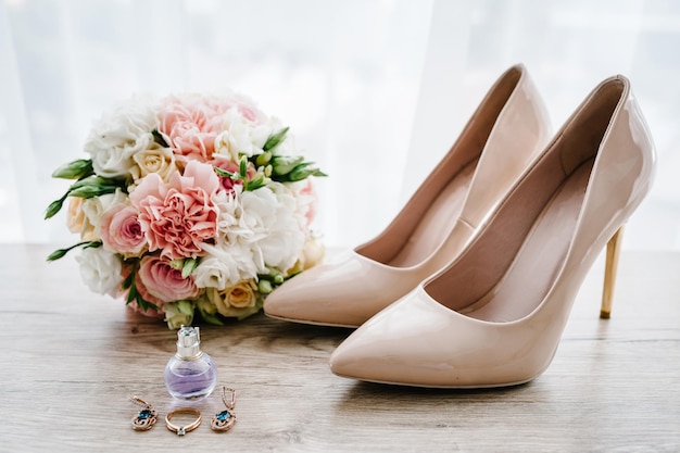 Wedding bouquet of the bride of pink flowers roses and greenery stylish classic lacquered beige shoes perfume earring and ring lying on wooden background Bride accessories Close up Side view