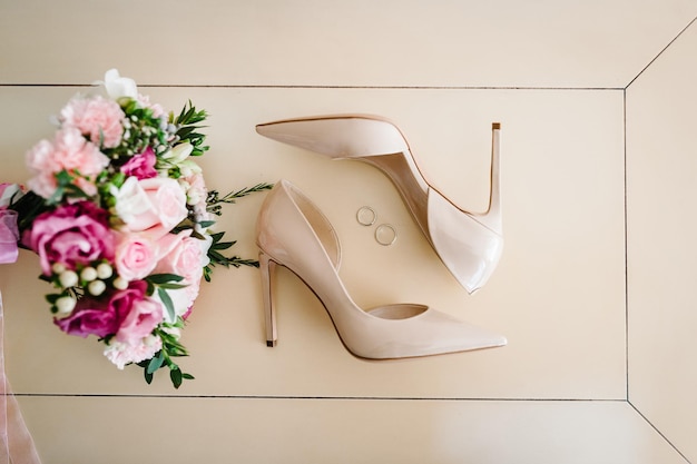 분홍색 꽃 장미와 녹색의 신부의 웨딩 부케는 파스텔 배경에 누워 있는 세련된 우아한 클래식 옻칠한 베이지색 신발과 2개의 은색 결혼 반지입니다.