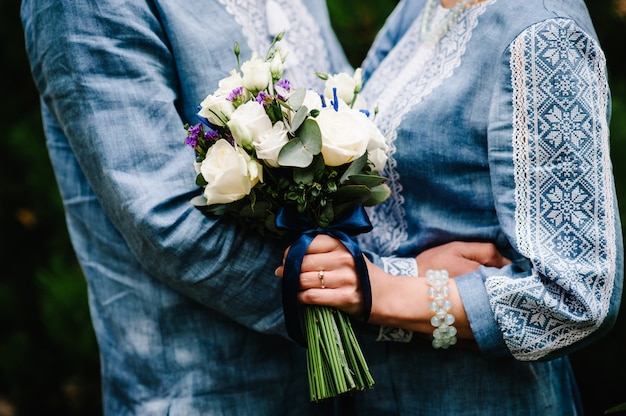 Wedding bouquet flowers from bush roses, eustoma in hands,  stylish brides woman wearing embroidered dress and groom in shirt holds a bouquet . Wedding ceremony. Close up.