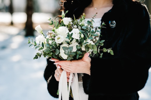 Wedding bouquet from white flowers and greenery in bride's hands on the background winter.
