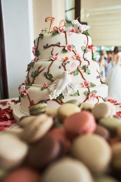 Wedding cake with decorative flowers, macarons, red roses petals and other different sweets on candy bar.