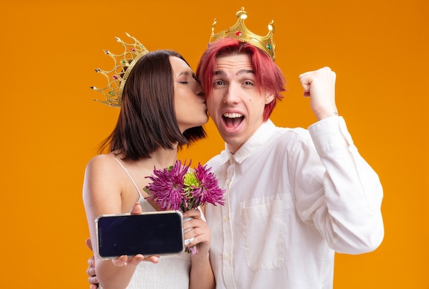 Wedding couple groom and bride with bouquet of flowers in wedding dress wearing gold crowns holding smartphone, groom happy and excited clenching fist