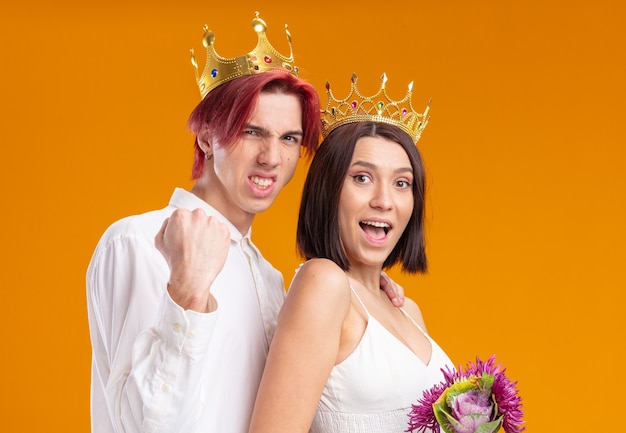 Wedding couple groom and bride with bouquet of flowers in wedding dress wearing gold crowns smiling cheerfully posing together clenching fist happy and excited