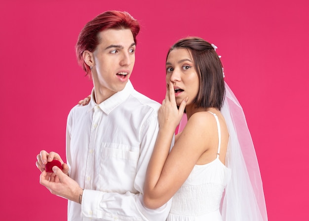 ピンクの壁にポーズをとって結婚指輪と赤いボックスを保持している白いドレスと新郎の結婚式のカップル幸せで興奮した花嫁