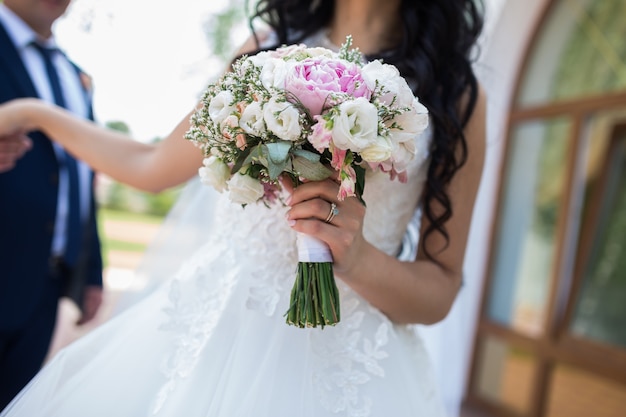 ピンクの牡丹とライラックユーストマのバラの花の豊富な束と夏の緑の背景に手をつないでいる結婚式のカップル。花束と結婚式のコンセプト