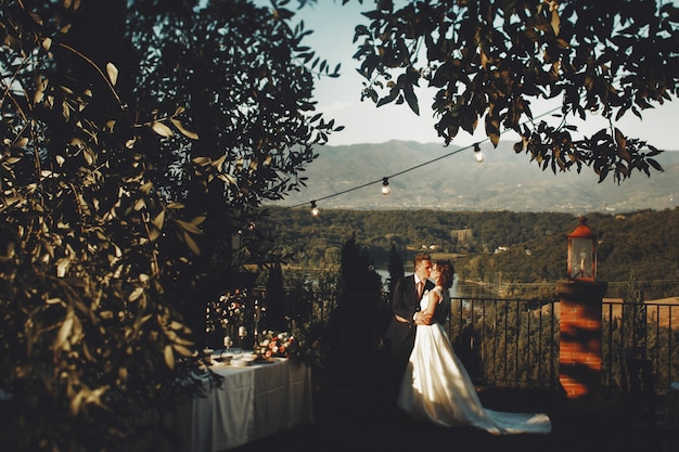 Photo wedding couple kisses standing on the porch with dinner table and evening lights