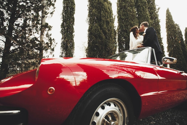 wedding couple kissing in a red cabrio which stands between tall trees